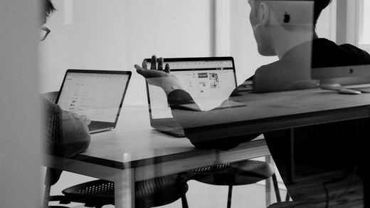 Two professionals collaborating and working on laptops viewed through a glass wall.
