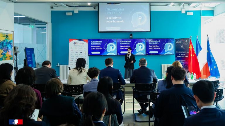 Audience attending a business conference in a modern event space, facing a stage with a large blue presentation screen and speakers. Participants are seated and standing throughout the room during a professional networking and knowledge-sharing event.