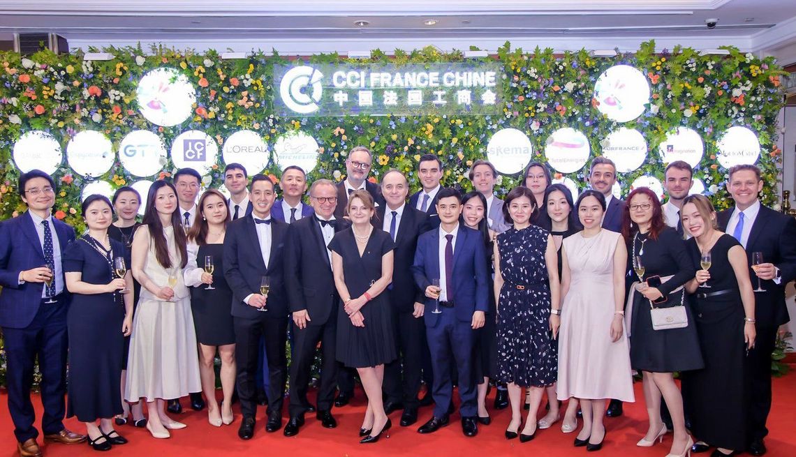 Large group of professionals posing for a photo on a red carpet in front of a CCI FRANCE CHINE flower backdrop with sponsor logos. Some attendees are holding champagne glasses.